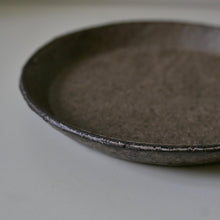 Load image into Gallery viewer, A close-up of a shallow round ceramic plate with a rustic, textured matte finish in dark gray-brown tones, set against a plain light background.