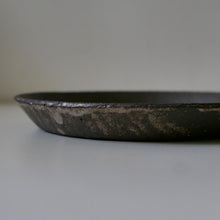 Load image into Gallery viewer, A close-up side view of a dark, rustic ceramic plate or shallow dish with a textured, distressed glaze showing light speckles and wear on a neutral gray background.

